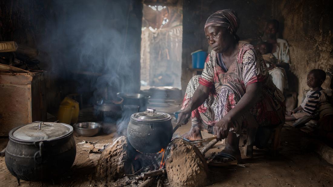 A woman cooking over a smoky open fire indoors with children nearby